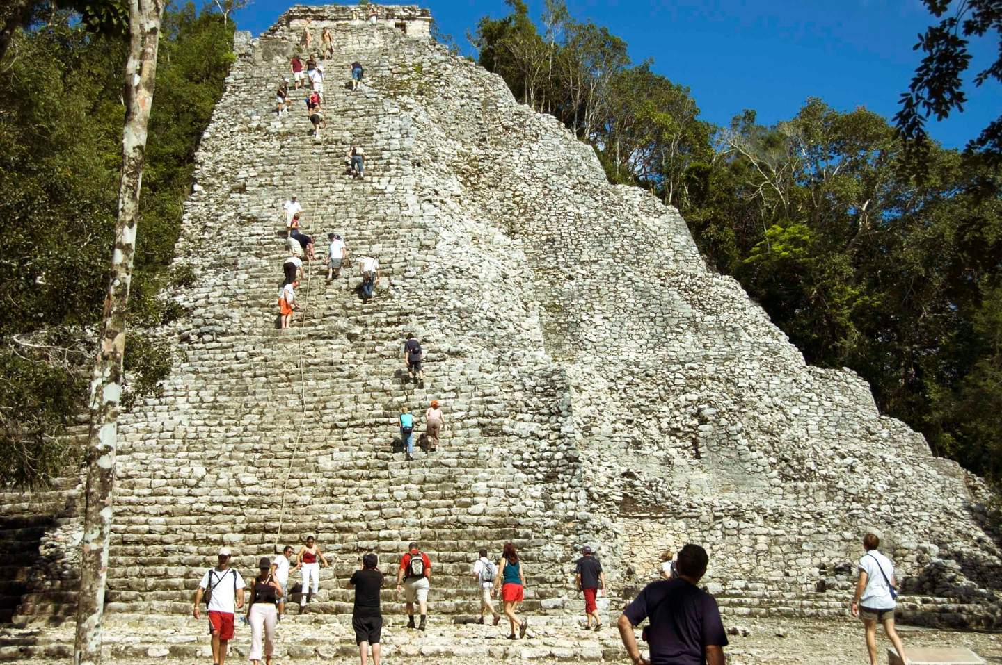 La gente escala una antigua pirámide de piedra en un entorno selvático en un día despejado, y muchos utilizan una cuerda para ayudarse.