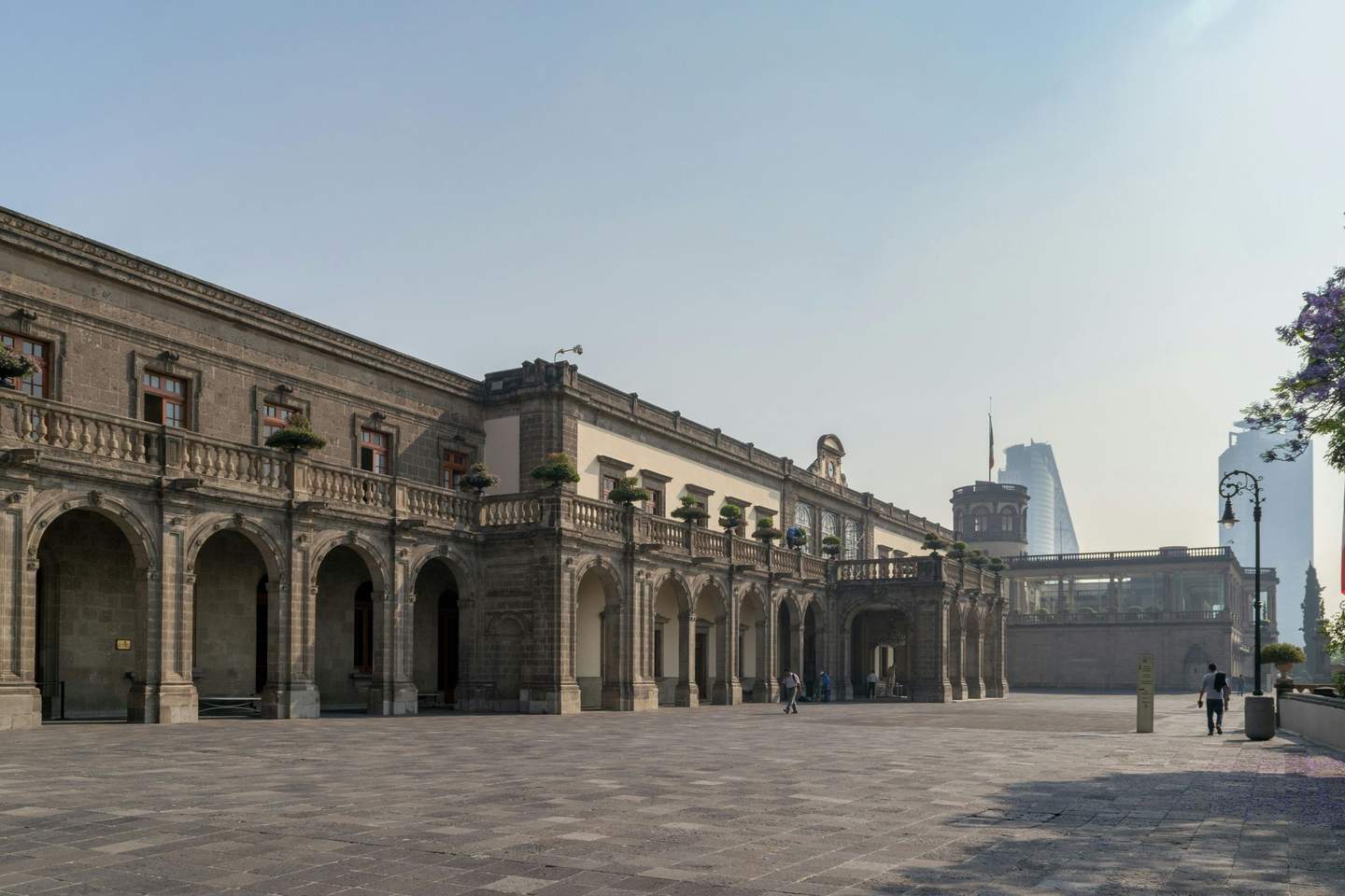 Historic stone building with arched colonnade and potted plants, spacious courtyard, few people walking, modern skyscraper in the distance.