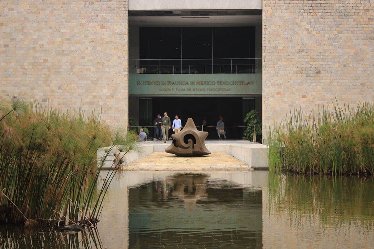 A stone building entrance with a large sculpture, a reflecting pool, and reeds; people are walking near the entrance.