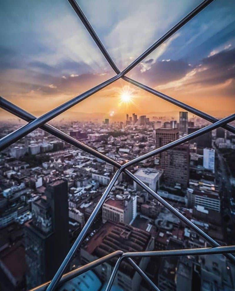 A cityscape at sunset viewed through a lattice of metal bars, with rays of sunlight illuminating the sky and buildings below.