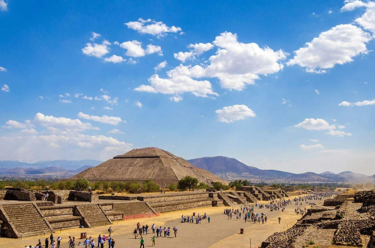 Teotihuacán Crowds of people visit ancient stone pyramids and structures under a bright, partially cloudy sky, with mountains in the background.