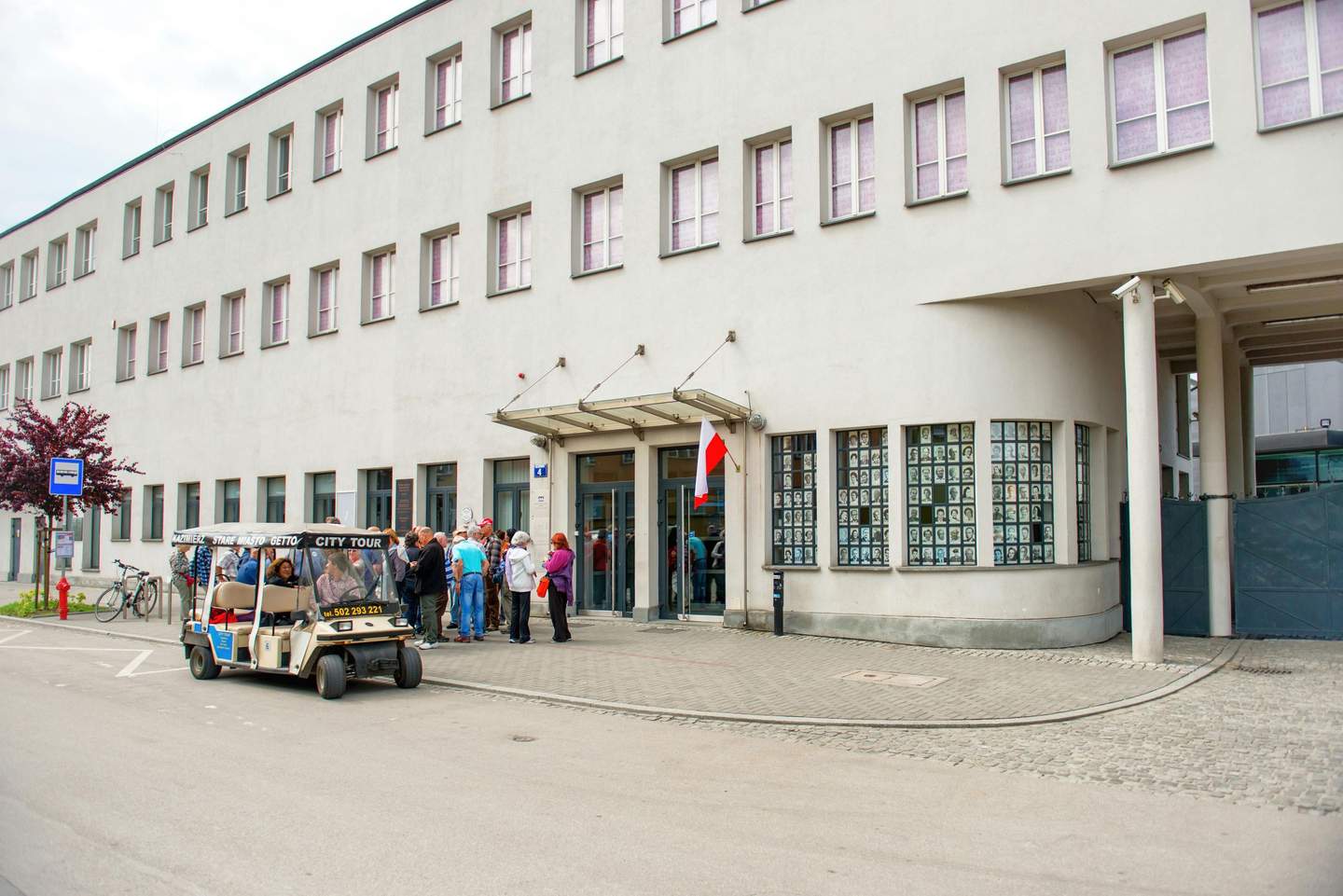 Oskar Schindler's Enamel Factory A group stands outside a building with displayed photos, next to a small vehicle labeled "City Tour" parked on the street.