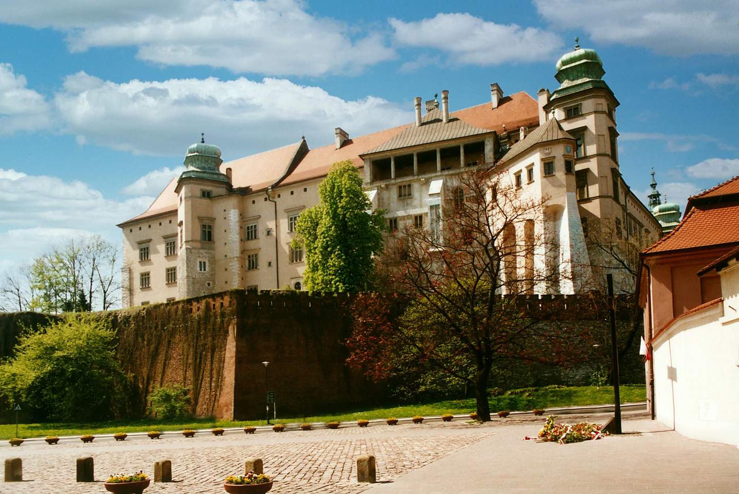 A large castle with green domes and red roofs stands atop a brick wall, surrounded by trees under a partly cloudy sky.