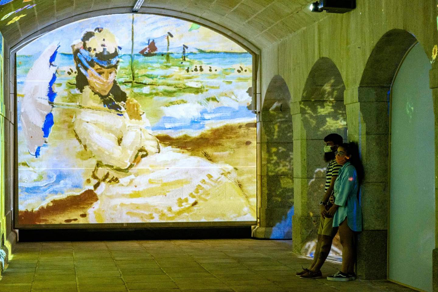 Immersivus Gallery Lisbon Two people stand in an arched hall with a large, colorful beach painting projected on the wall.