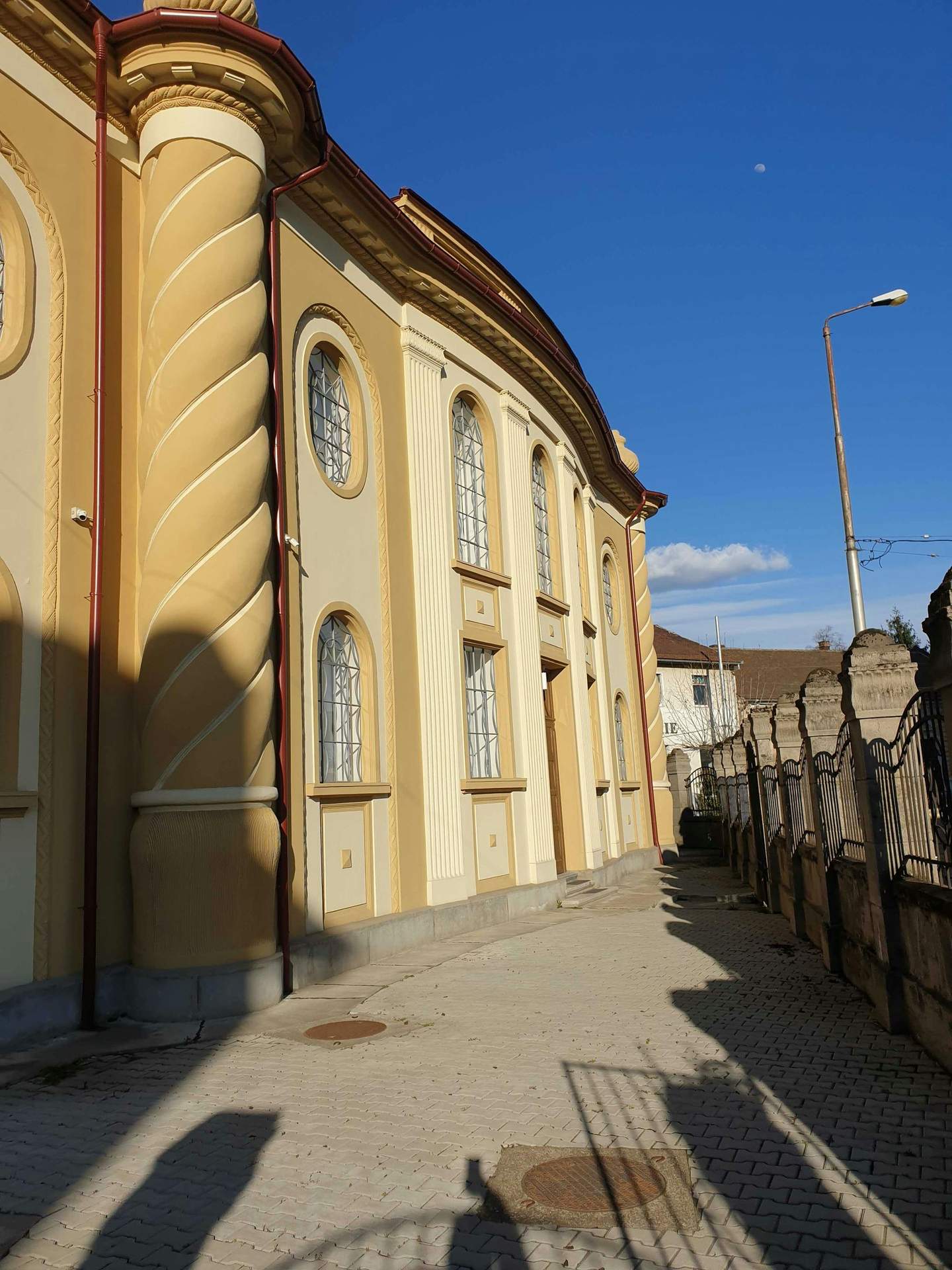 Museum of Jewish History in Oradea Yellow building with detailed columns and arched windows adjacent to a paved walkway and ornate iron fencing under a blue sky.