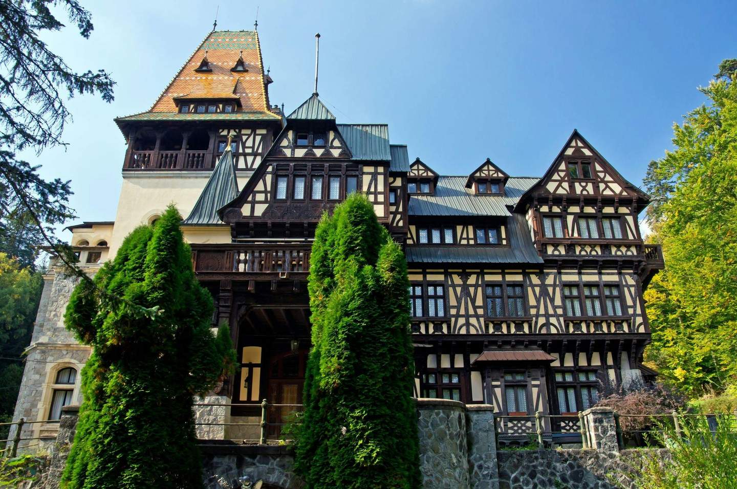 Pelișor Castle Large, ornate Tudoresque building with intricate wood detailing and tall, peaked roof surrounded by trees under a clear blue sky.