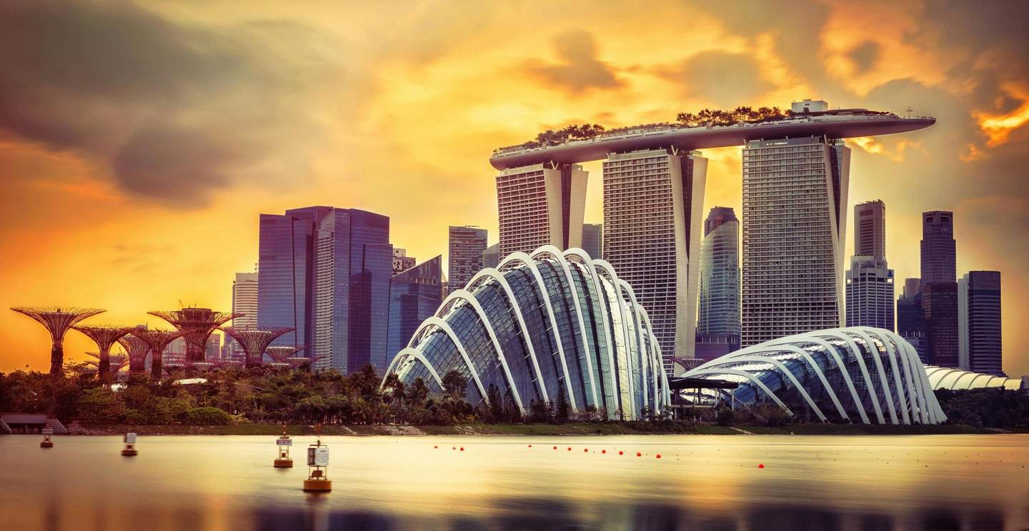 Paisagem urbana com edifícios futuristas, incluindo o Marina Bay Sands e o Supertree Grove, ao pôr do sol, com água em primeiro plano.