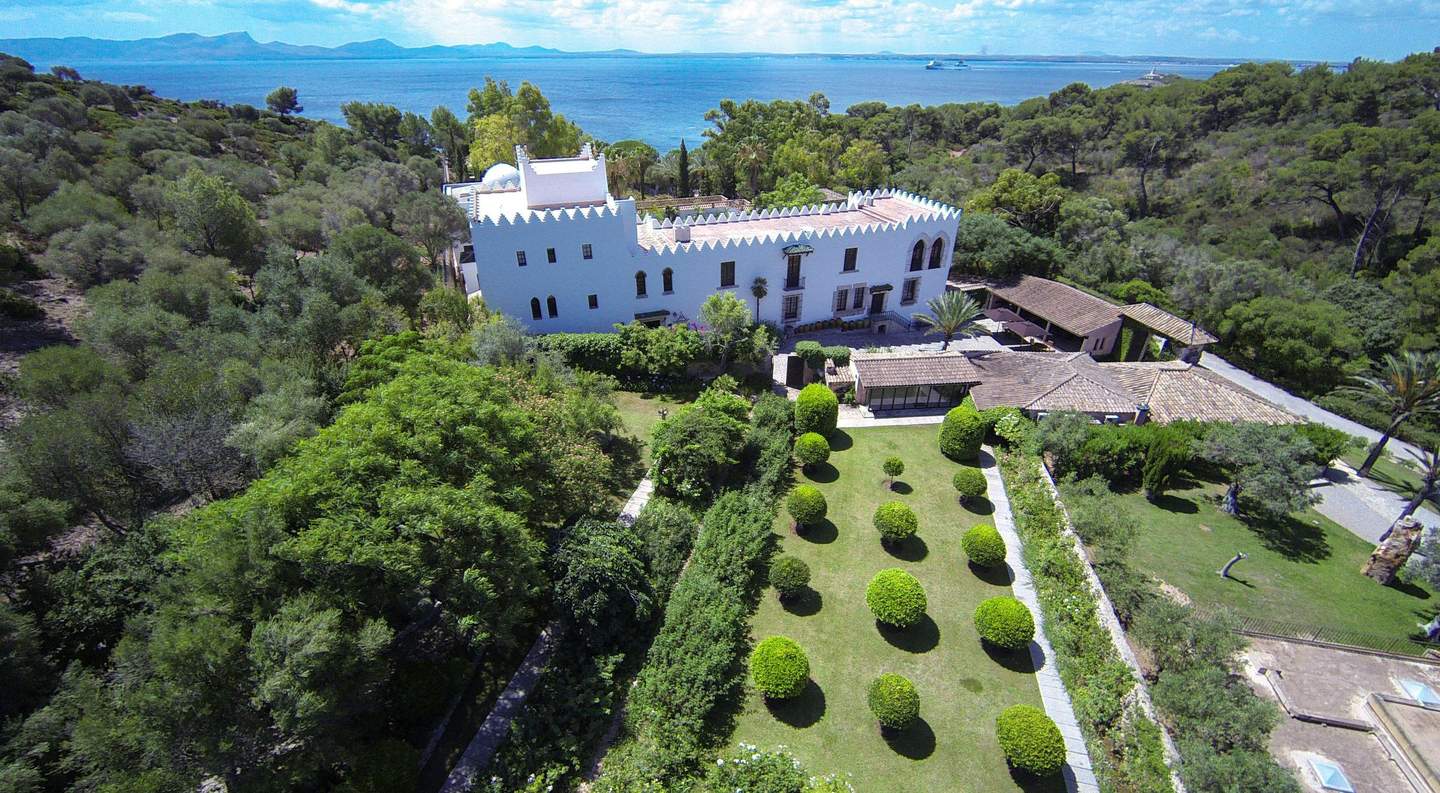 Aerial view of a large white building with battlements, surrounded by lush gardens, trees, and adjacent coastal waters.