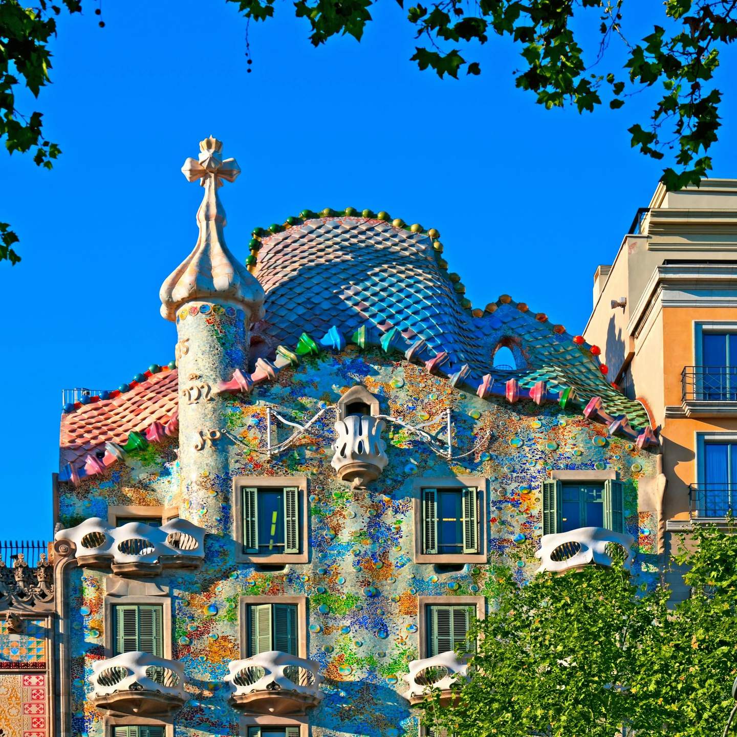 Casa Batlló A colorful, mosaic-covered building with curved balconies, green shutters, and a cone-shaped turret, framed by tree branches.