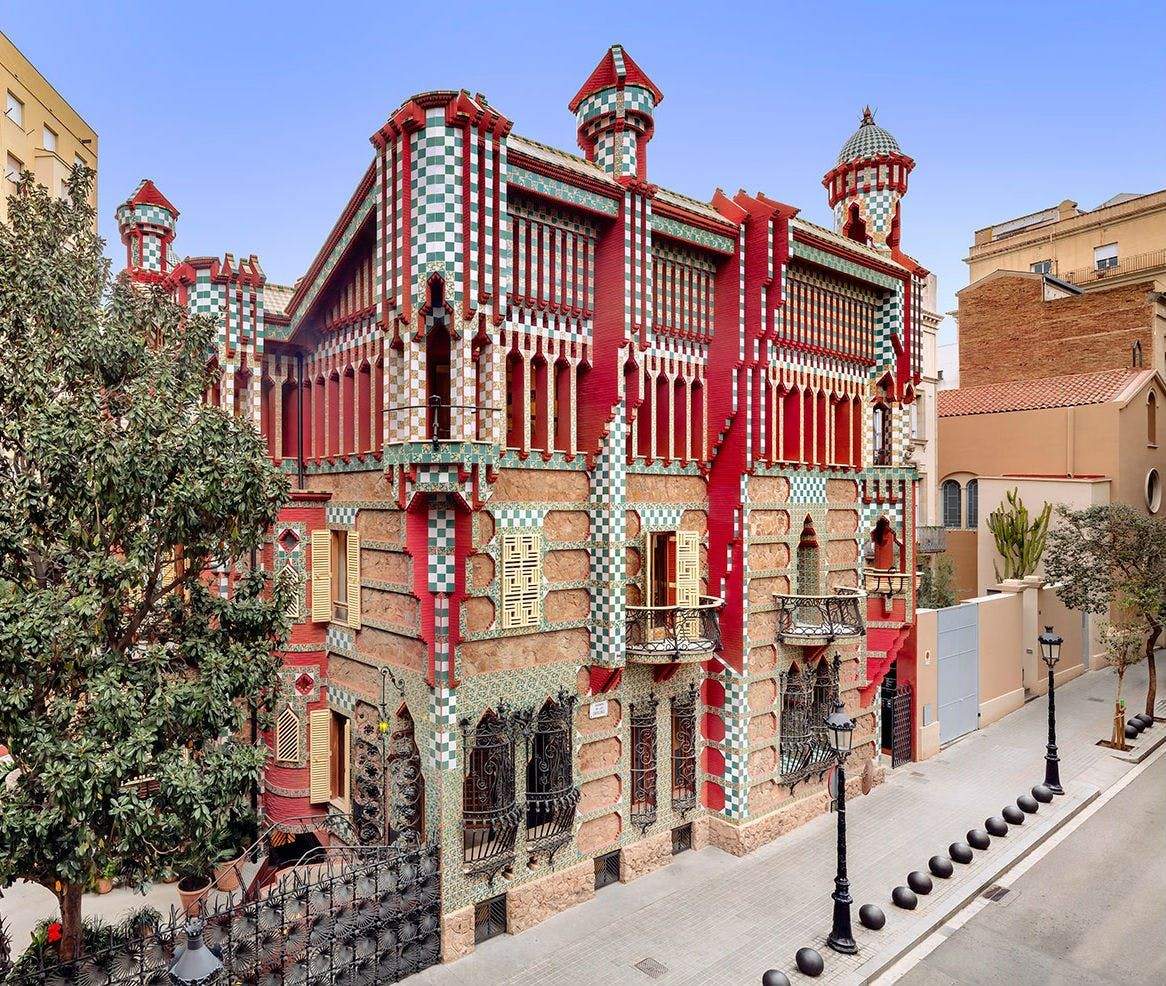 A vibrantly decorated building with checkered patterns, red accents, and intricate ironwork on a quiet street lined with trees and lamps.