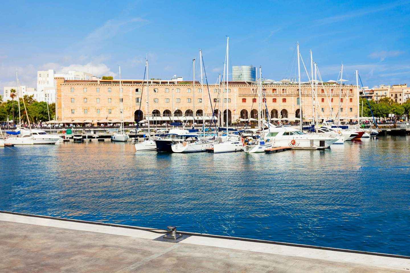 Museu d'Història de Catalunya Boats and yachts docked in a marina with a large, arched-windowed building and clear blue sky in the background.
