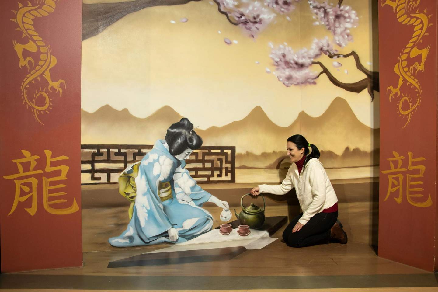 Museum of Illusions Barcelona A woman interacts with a mural depicting a traditional Japanese tea scene against a backdrop of mountains and cherry blossoms.