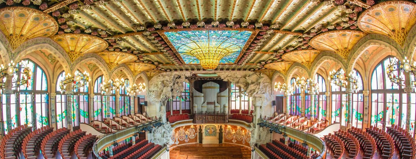 Palau de la Música Catalana Ornate concert hall with arched stained glass windows, intricate ceiling designs, chandeliers, and rows of red seats.