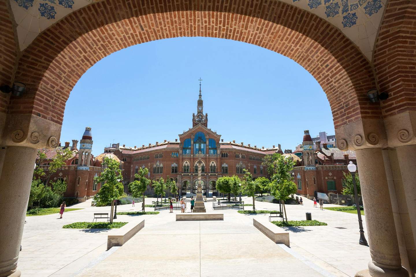 People walk in a courtyard framed by an archway, with a large, ornate brick building in the background under a clear blue sky.