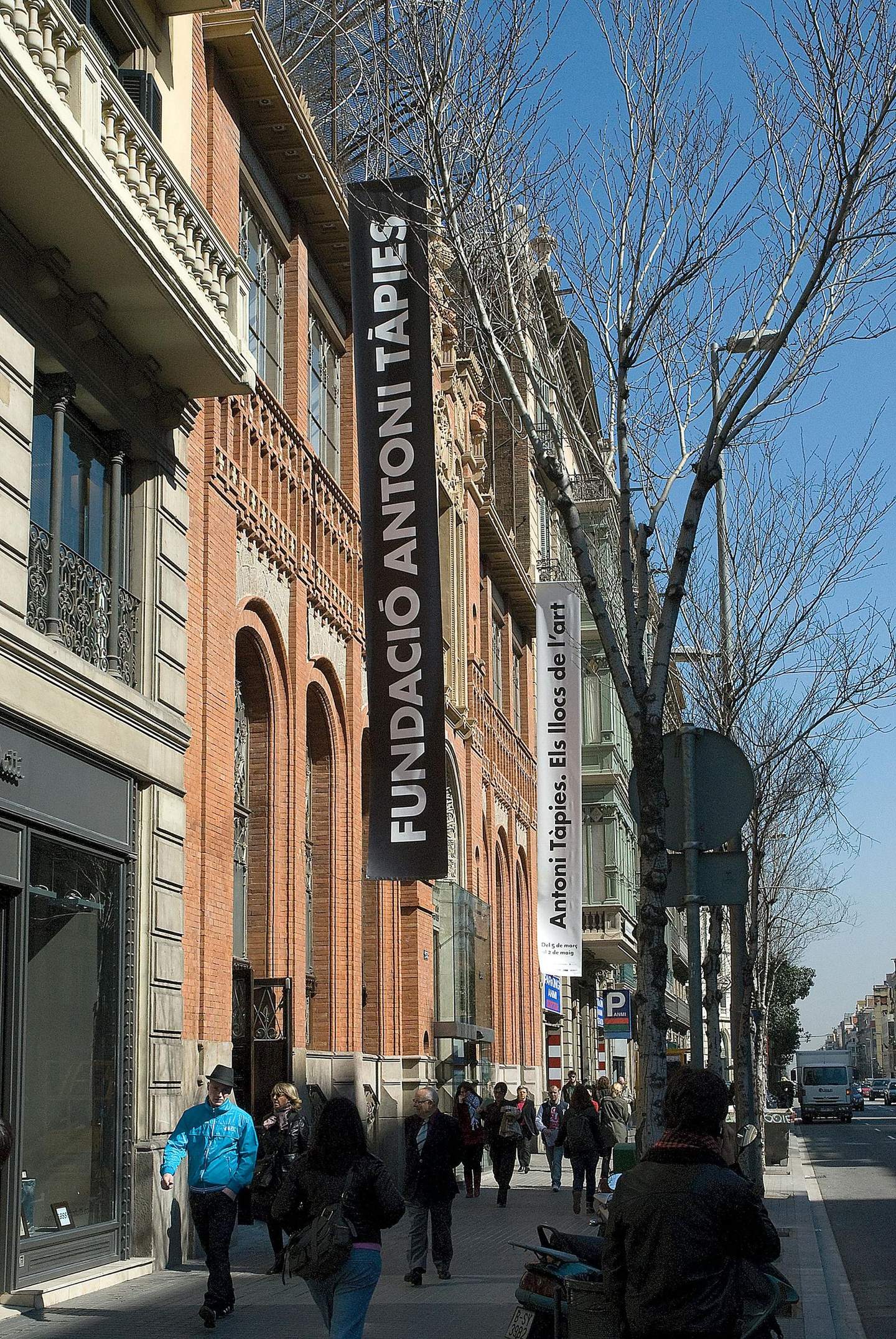Tàpies Museum People walk by the Fundació Antoni Tàpies building on a bustling street lined with trees and various storefronts.