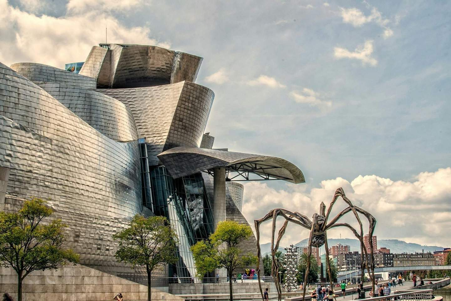 Guggenheim Museum Bilbao Modern building with curving metal surfaces, large abstract spider sculpture, trees, and people walking on a sunny day.
