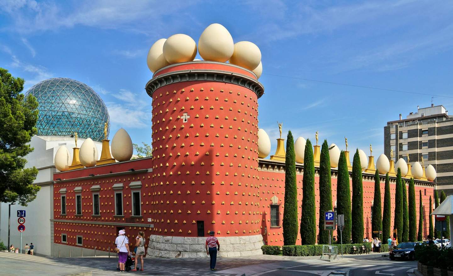 Dalí Theatre and Museum Red building with large decorative eggs on the roof, dotted with gold, and a glass dome in the background. People walking nearby.