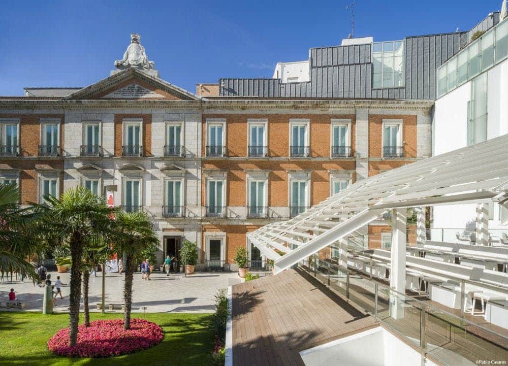 Museo Nacional Thyssen-Bornemisza Courtyard with palm trees and pink flowers, adjacent to a multi-story building with balconies and a modern glass structure.
