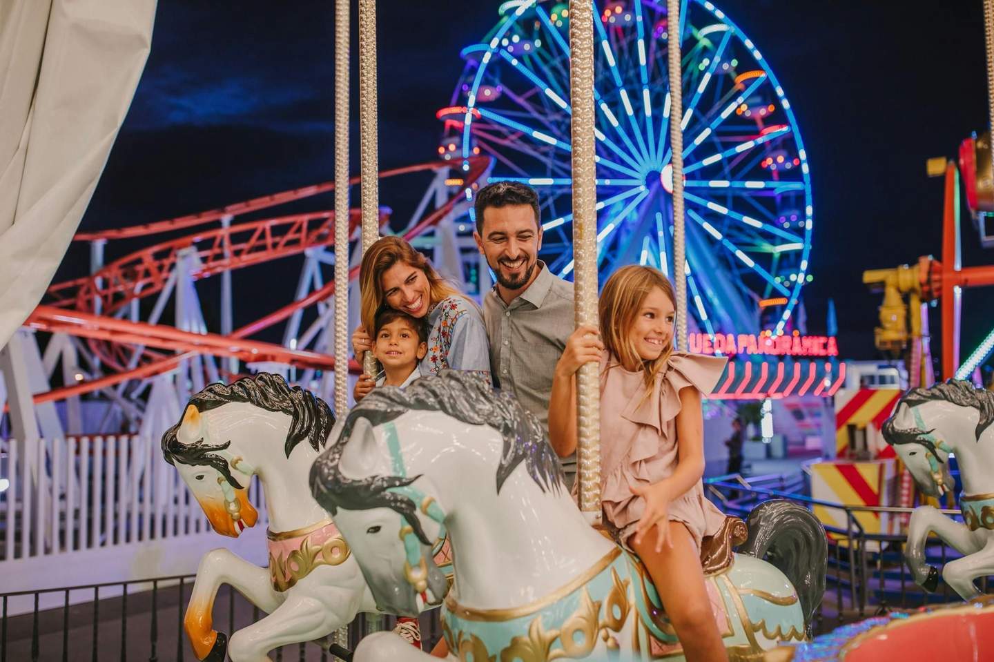 Holidayworld Maspalomas Center A happy family rides a carousel at night with a lit Ferris wheel and roller coaster in the background.