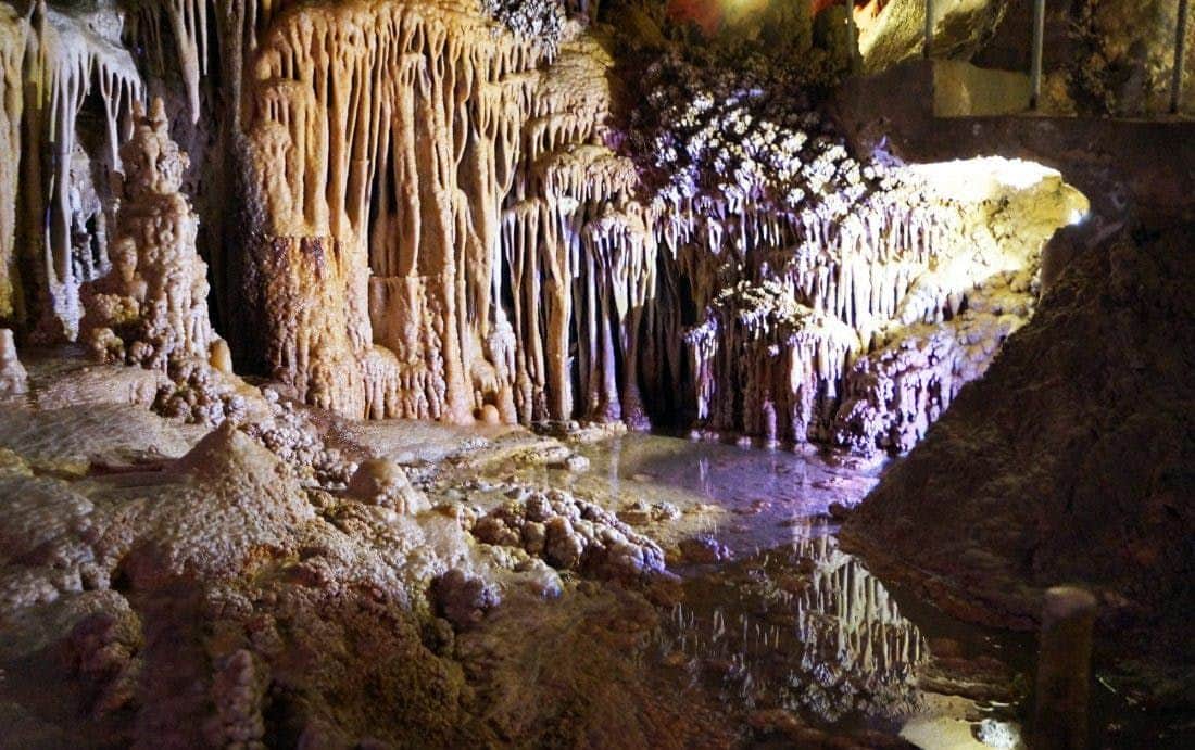Genova Caves A cave interior with formations of stalactites and stalagmites, a small pool of water reflecting light, and rocks covered in minerals.