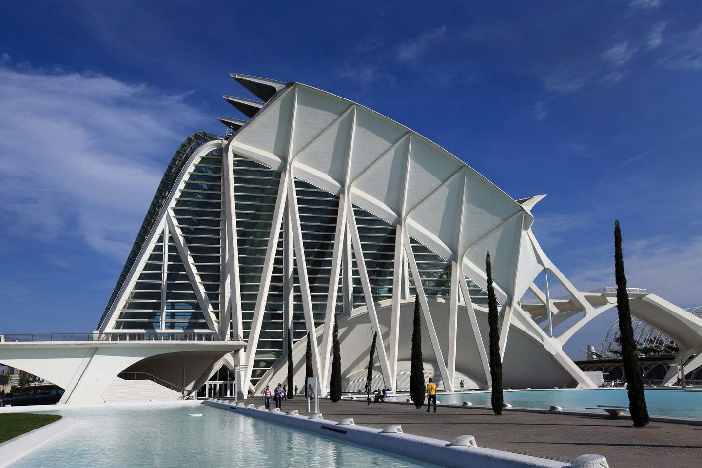 A futuristic white building with structural arches, surrounded by water and a few tall, narrow trees against a blue sky.