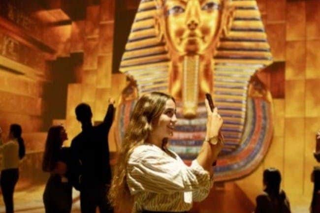 Tutankhamun Exhibition Stockholm A woman takes a photo with her phone in front of a large illuminated display of a golden pharaoh mask in a museum-like setting.
