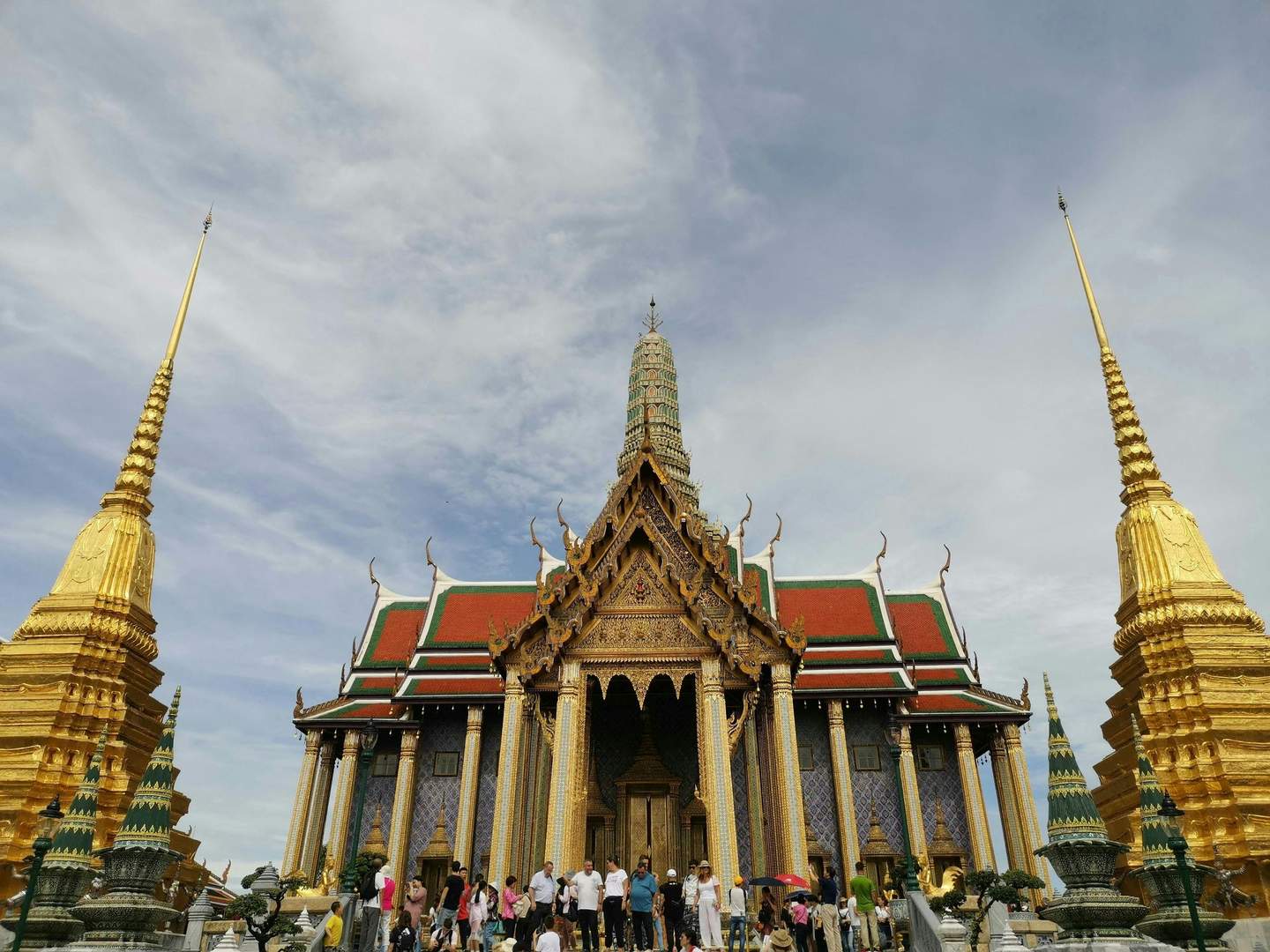 The Temple of the Emerald Buddha Tourists visit an ornate temple with golden spires, intricate details, and colorful rooftops under a partly cloudy sky.