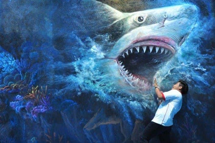 A person poses in front of a mural depicting a large shark emerging from the water with its mouth open.