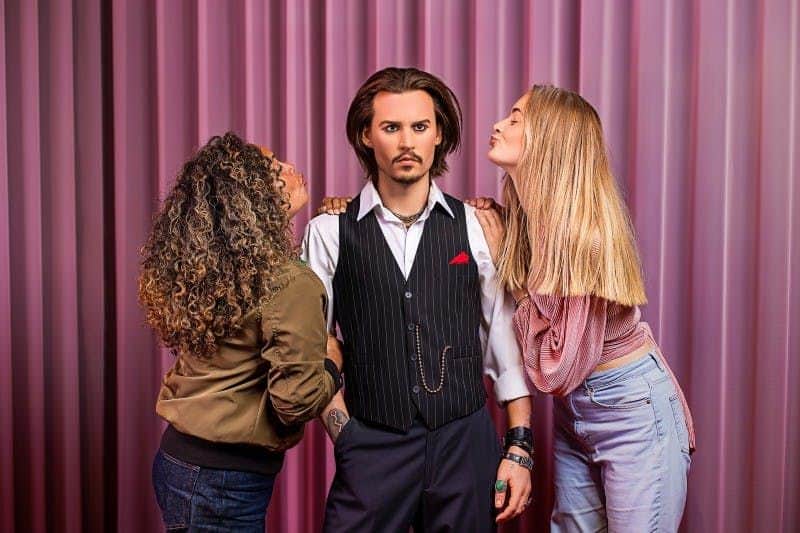 Three people pose in front of a pink curtain; a man in a vest stands between two women, one on each side, both leaning in toward him.