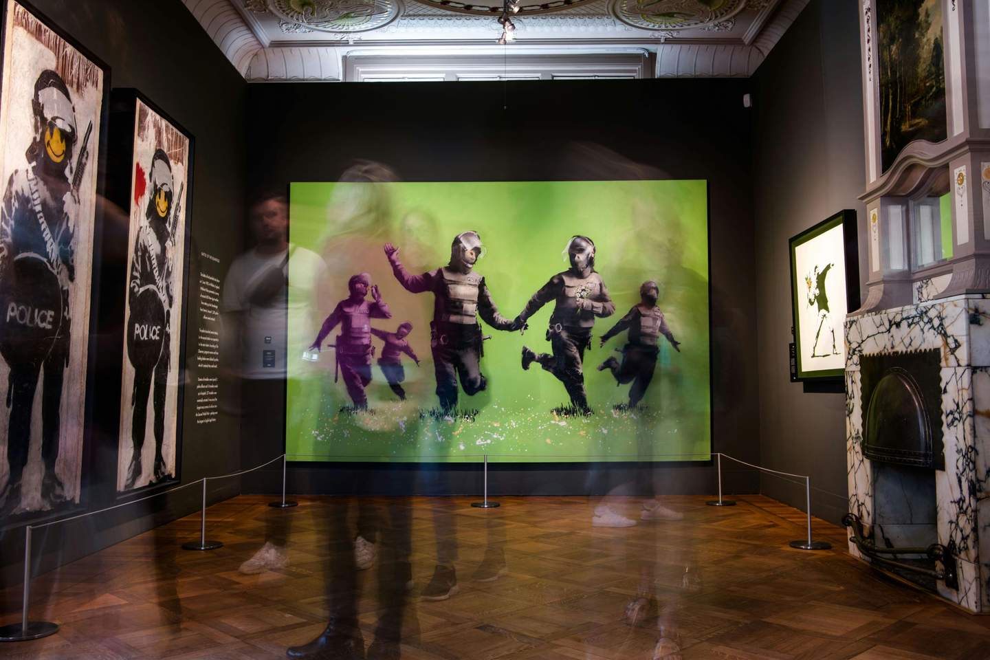 Moco Museum Amsterdam Art gallery with blurred motion of people viewing Banksy artworks, including a large green mural of police officers and smaller framed pieces.