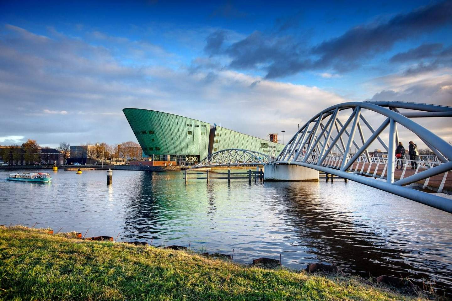 NEMO Science Museum Futuristic green building near a waterfront with a distinctive bridge and a passing boat under a partly cloudy blue sky.