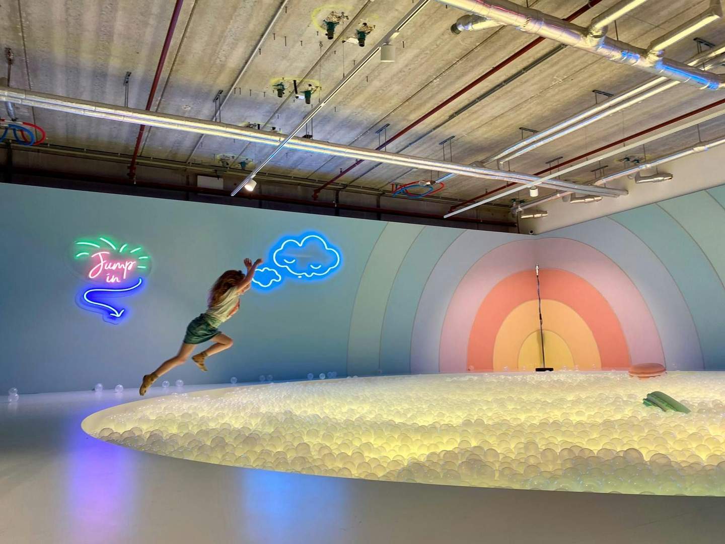The Upside Down Person jumping into an illuminated ball pit with neon signs, rainbow mural, and industrial ceiling pipes in the background.