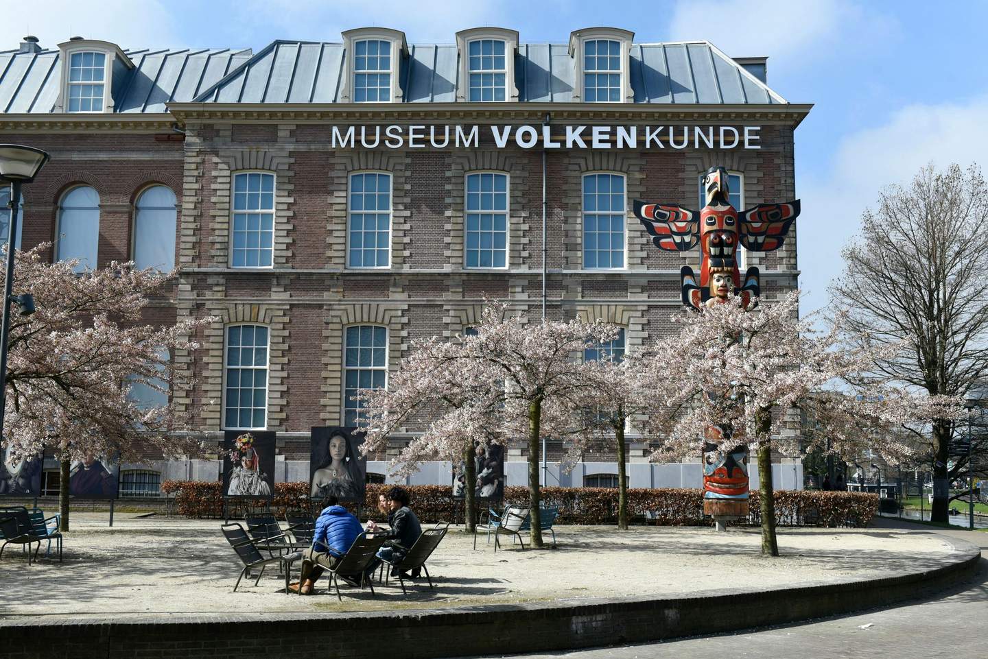 Wereldmuseum Leiden People sitting on benches under blooming cherry trees in front of a museum building with "MUSEUM VOLKENKUNDE" sign.