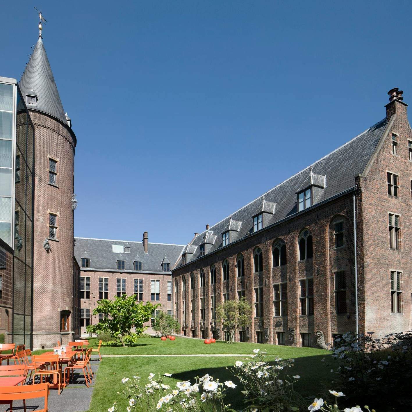 A historic brick building with a tower, windows, a lawn with orange chairs and tables, flowers, and red spheres under a clear blue sky.