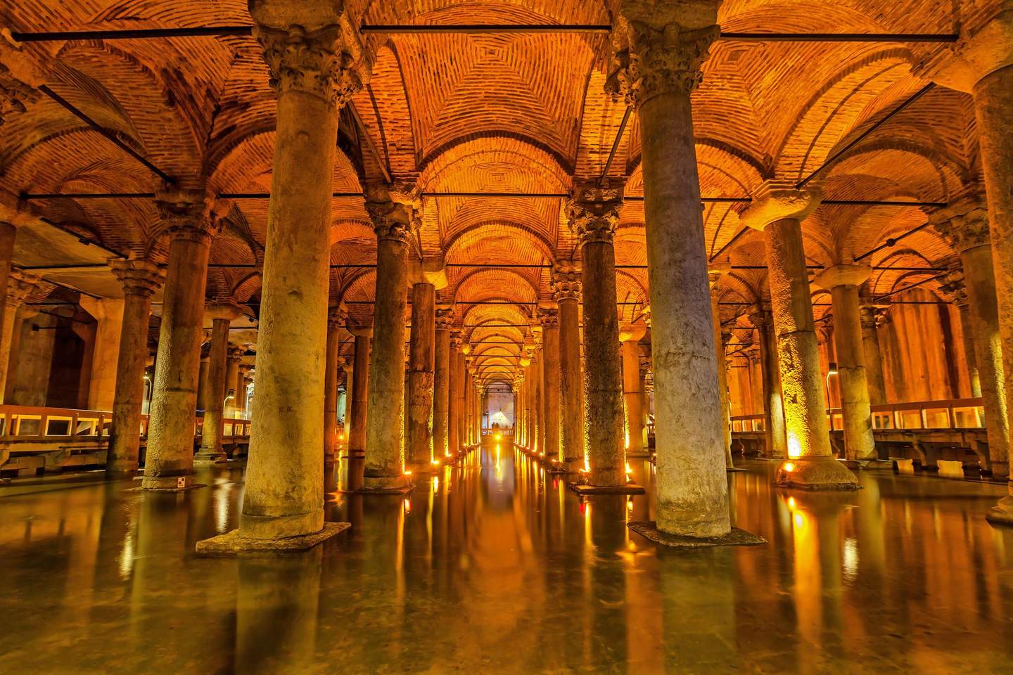 A cavernous subterranean chamber with numerous stone columns, arched brick ceilings, and dim, warm lighting reflecting on water.