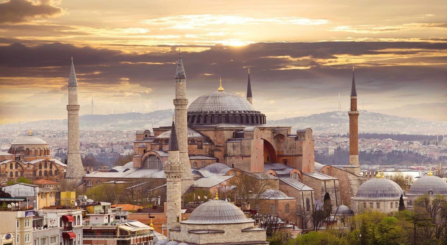 Hagia Sophia An ancient mosque with multiple minarets at sunset, situated in a cityscape with residential buildings and hills in the background.