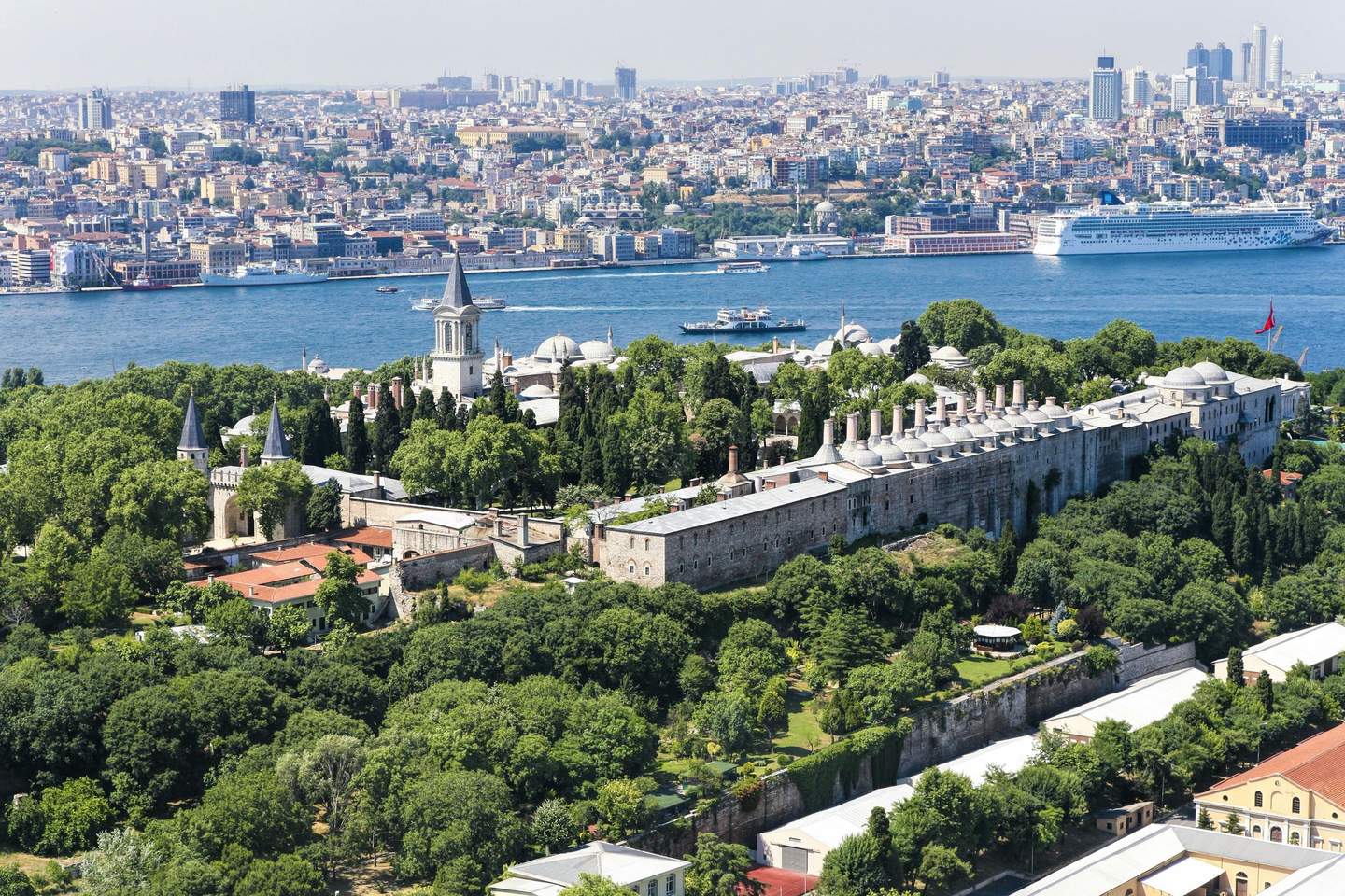 Historic palace complex with towers and domes surrounded by lush greenery, set near a river with a cityscape in the background.