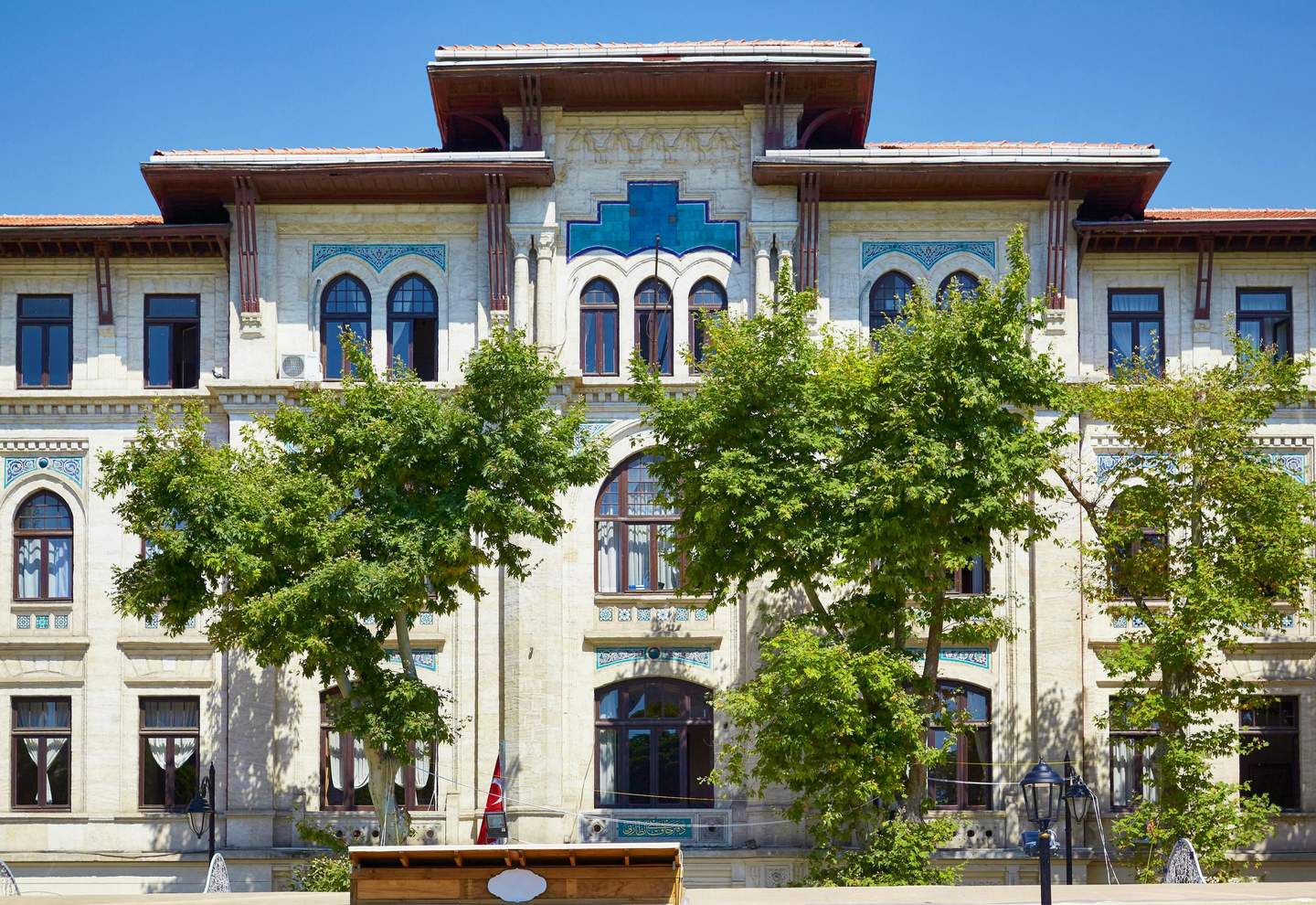 Turkish and Islamic Arts Museum Ornate stone building with large arched windows, teal tile details, and red roof, partially obscured by green trees under a clear blue sky.