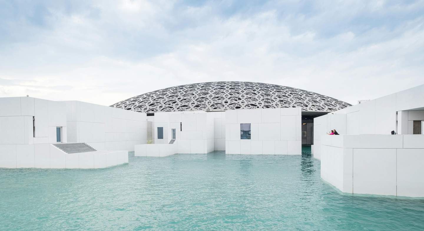 Louvre Abu Dhabi White geometric buildings surrounded by water, with an intricate dome structure in the background and a blue sky.