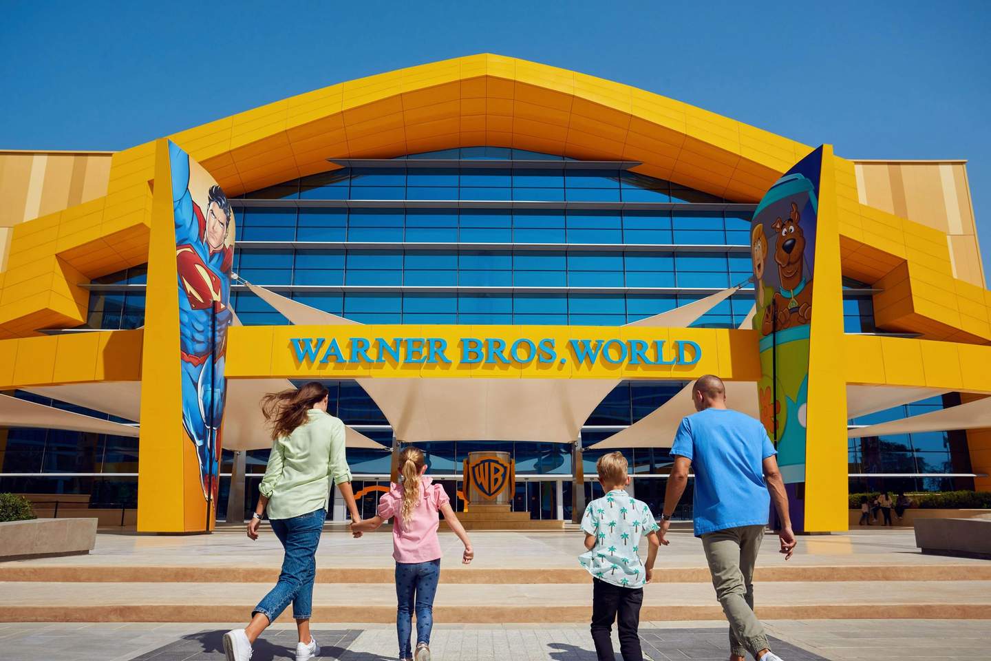 Warner Bros. World™ Abu Dhabi A family walking towards the entrance of Warner Bros. World, with Superman and Scooby-Doo artwork on the building's facade.