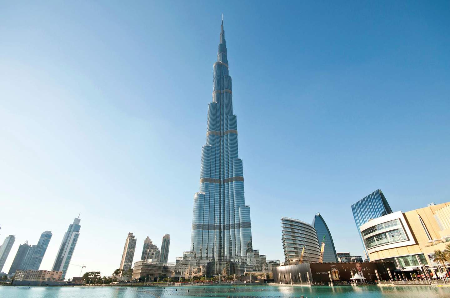 Tall skyscraper in a city with modern buildings and a reflecting pool in the foreground, under a clear blue sky.