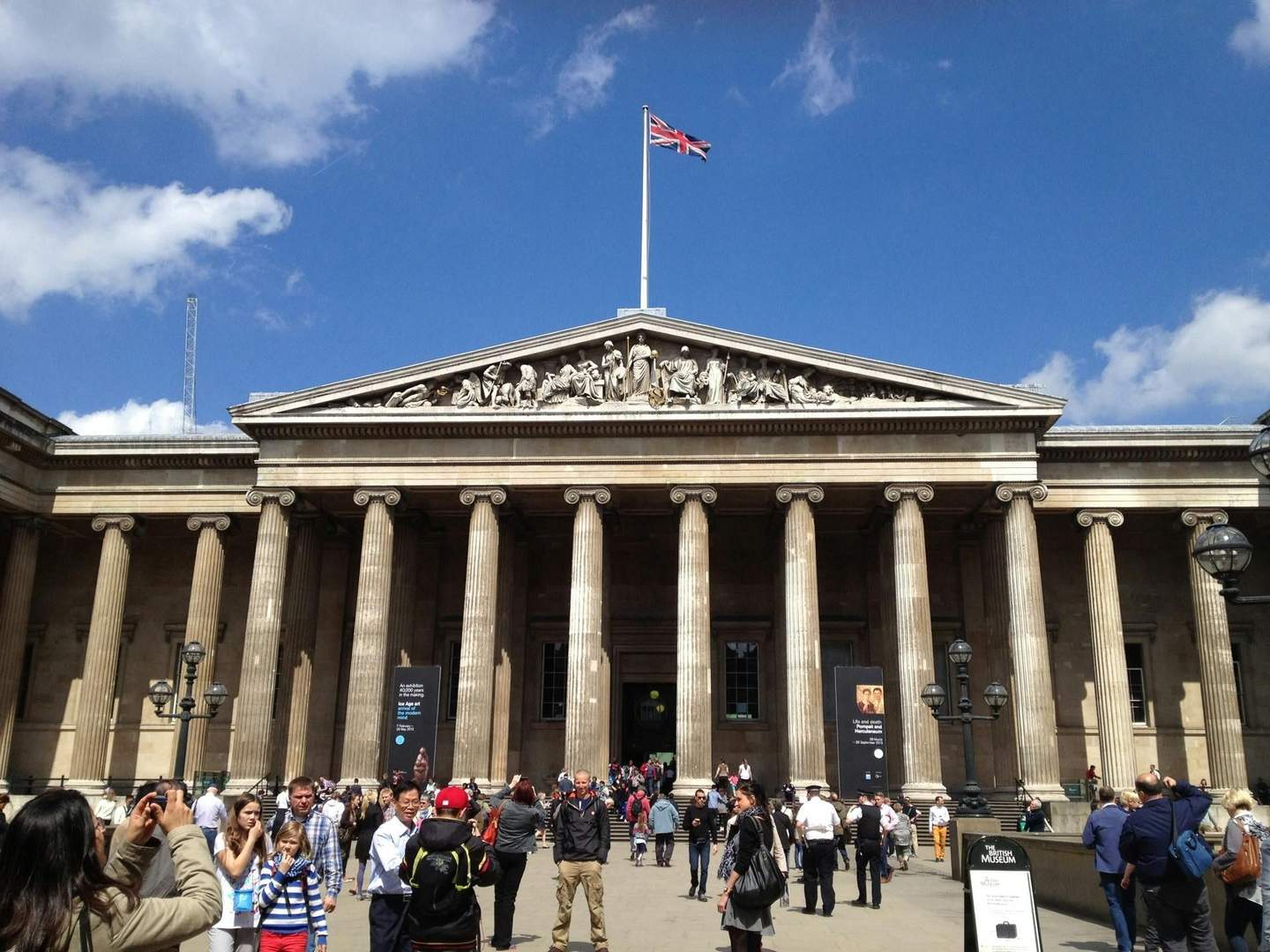 The British Museum A group of people are gathered in front of a building with columns and a British flag on top, under a blue sky.