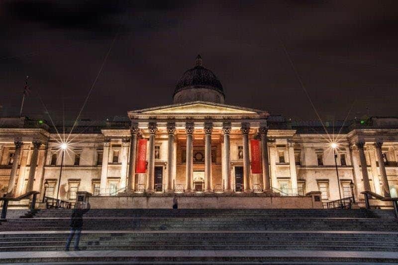The National Gallery Illuminated neoclassical building with columns and dome at night, with banners hanging and a person on the steps in the foreground.
