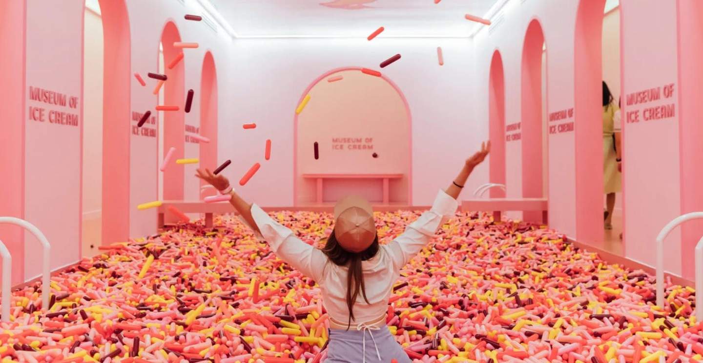 Person in a hat throws colorful plastic "sprinkles" in a pink-themed room labeled "Museum of Ice Cream."