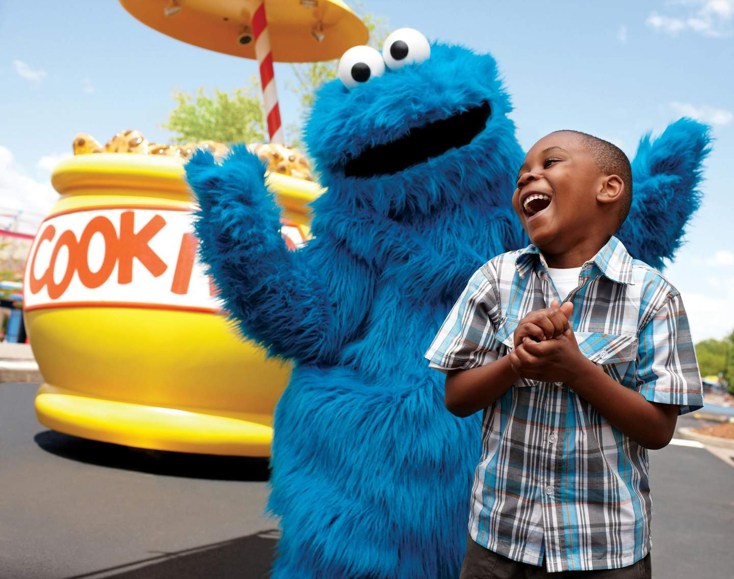 Sesame Place Child laughing with Cookie Monster mascot outdoors near large cookie jar prop.