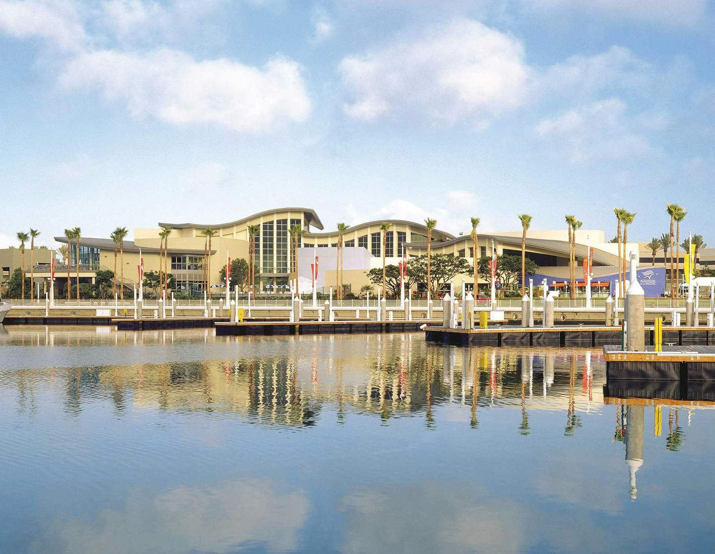 A waterside view of a modern building with palm trees, docks, and calm water reflecting the scene under a partly cloudy sky.