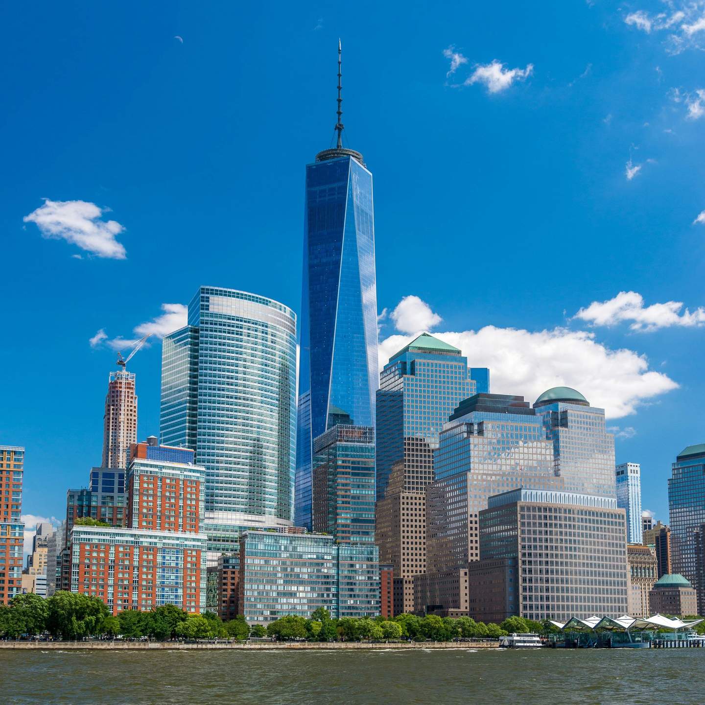 One World Observatory Several modern skyscrapers, including a tall, pointed tower, stand along the waterfront under a clear blue sky.
