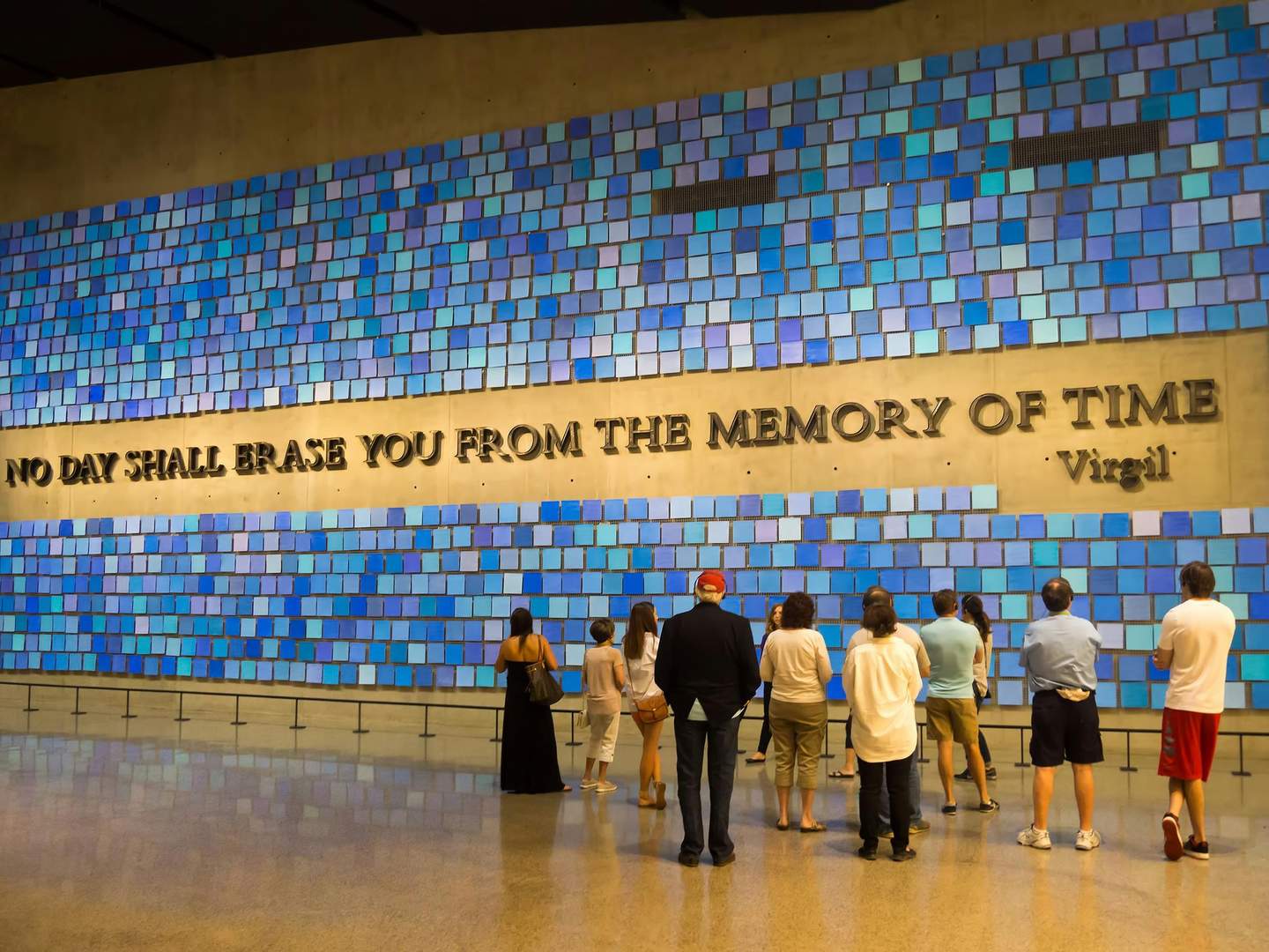 The 9/11 Memorial & Museum People standing before a wall of blue tiles with the inscription "NO DAY SHALL ERASE YOU FROM THE MEMORY OF TIME - Virgil."