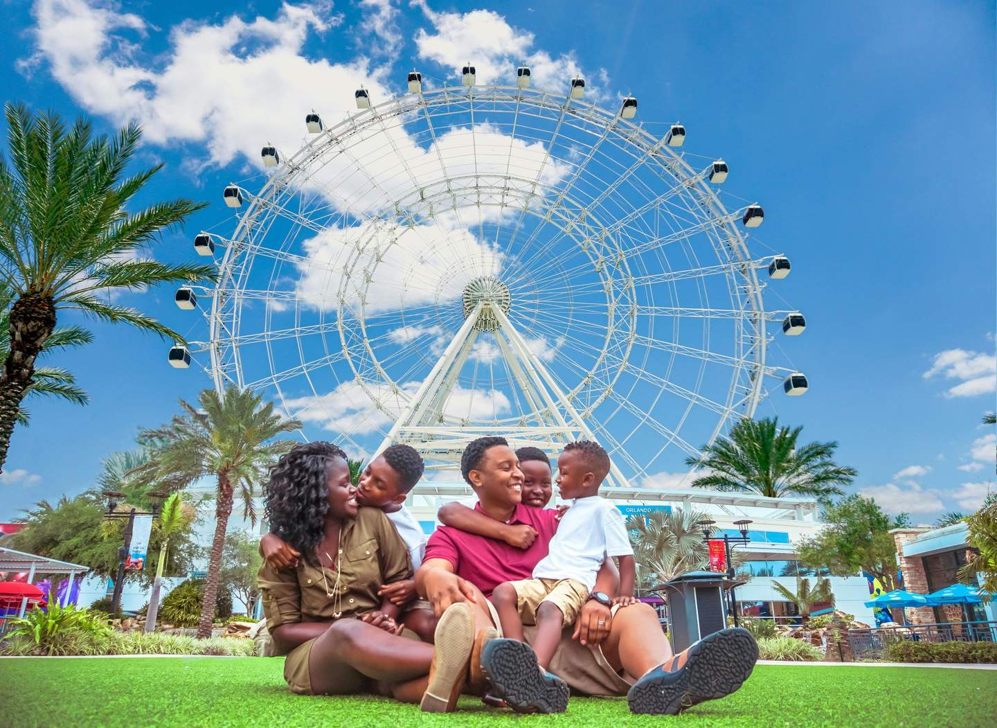 ICON Park™ A family of five sits on grass, smiling in front of a large Ferris wheel, with palm trees and a clear blue sky in the background.