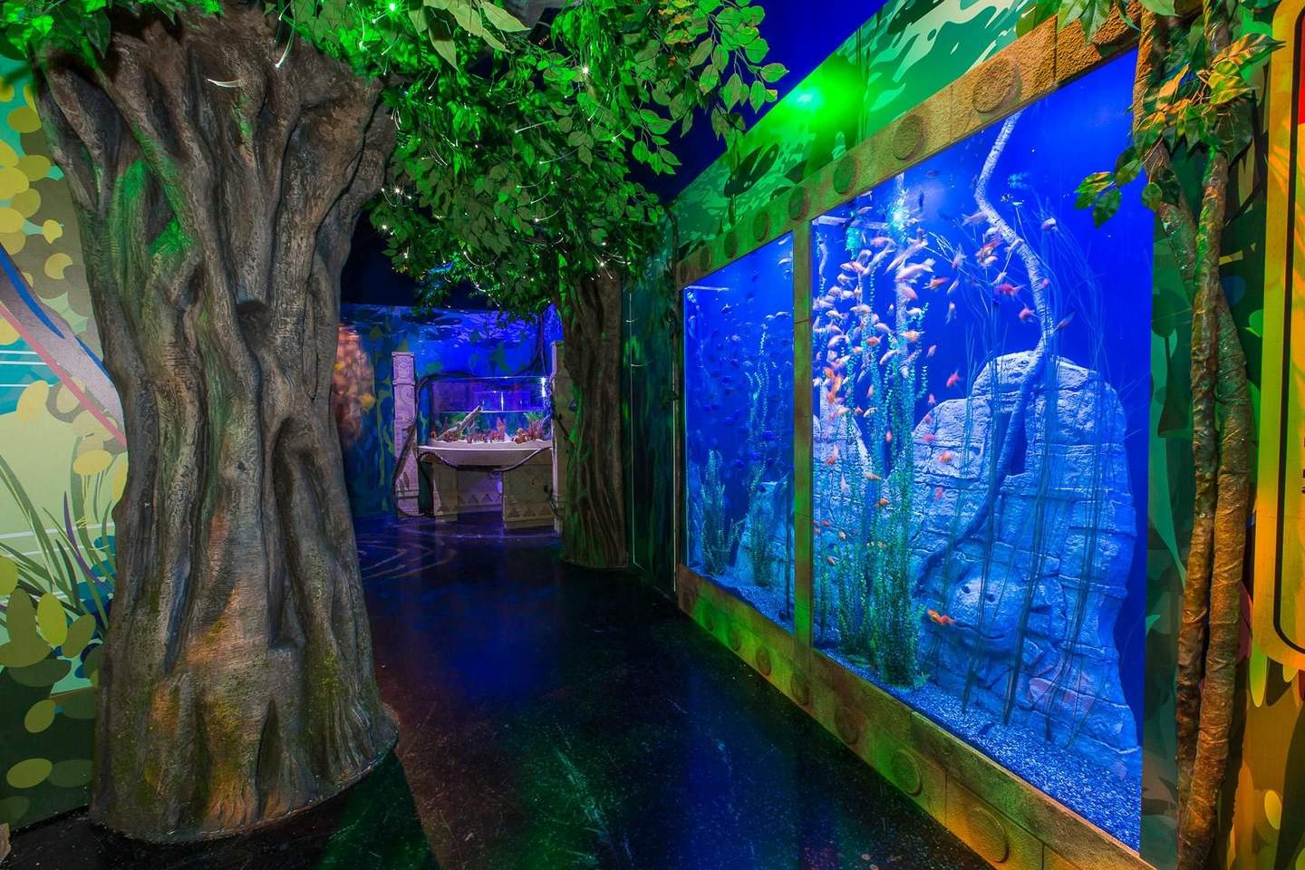 An indoor aquarium with large fish tanks, tree-like structures, lush green foliage, and vibrant lighting in a jungle-themed setting.